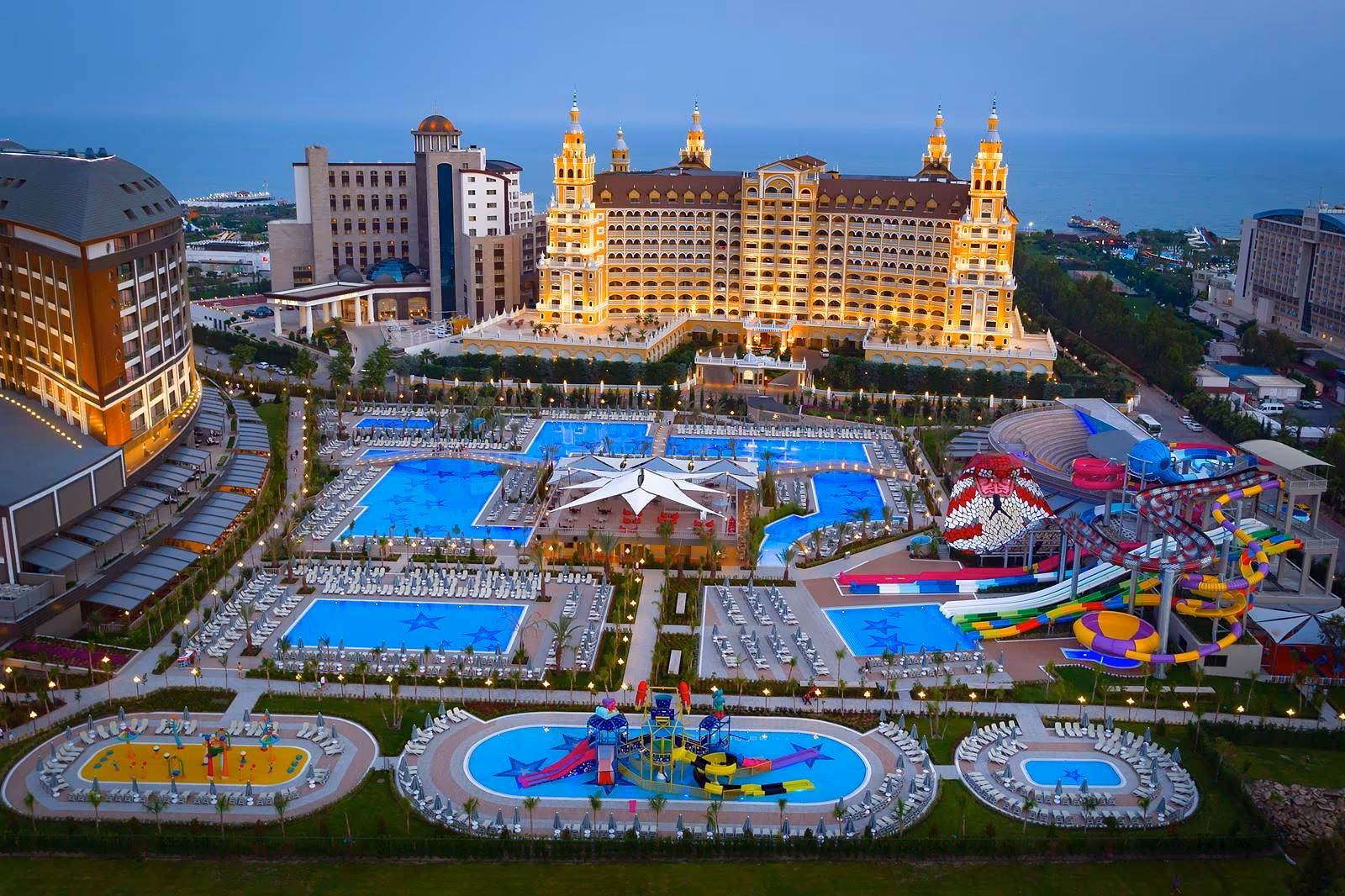 Luchtfoto in de schemering van het zwembadlandschap met een stervormig plein en het aquapark, met een goudkleurig hotel op de achtergrond bij Royal Seginus in Lara