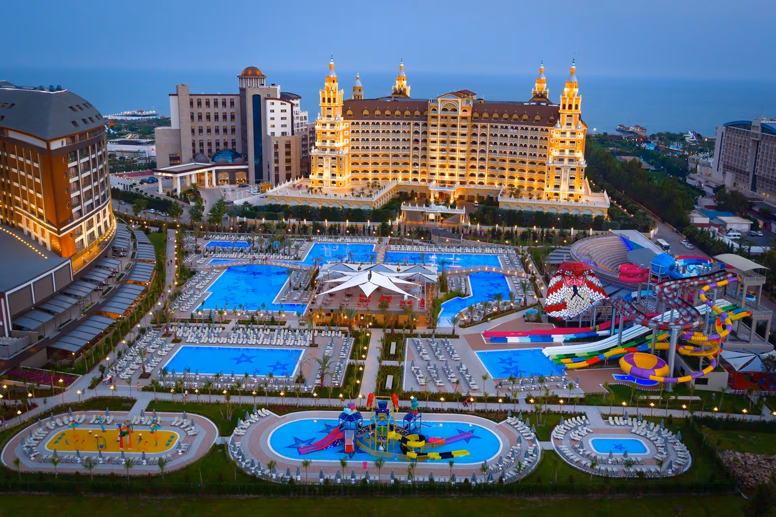 Luchtfoto in de schemering van het zwembadlandschap met een stervormig plein en het aquapark, met een goudkleurig hotel op de achtergrond bij Royal Seginus in Lara