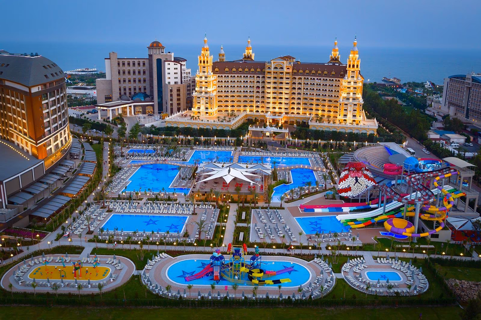 Luchtfoto in de schemering van het zwembadlandschap met een stervormig plein en het aquapark, met een goudkleurig hotel op de achtergrond bij Royal Seginus in Lara