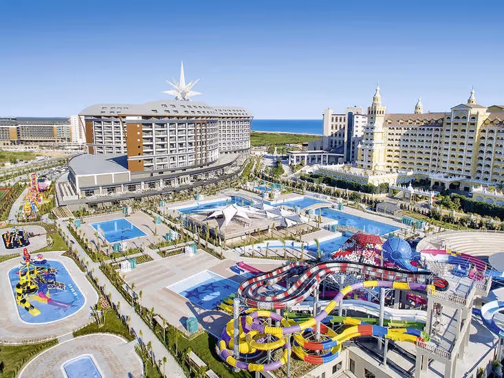 Luchtfoto van het hoofdgebouw, het aquapark met gekleurde glijbanen en de hotels ernaast, met de zee aan de horizon bij Royal Seginus in Lara